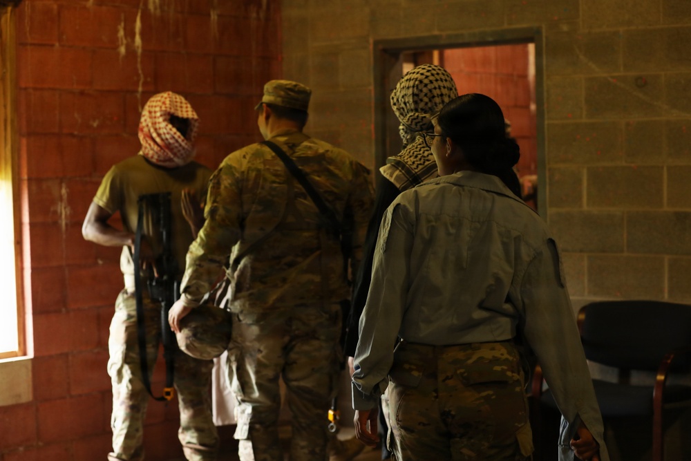 The image shows several individuals inside a brick building, viewed from behind. At least one person is wearing military camouflage uniform and a patrol cap, while others wear keffiyeh-style head coverings and mixed attire. The setting and equipment suggest a military training exercise or role-player scenario.