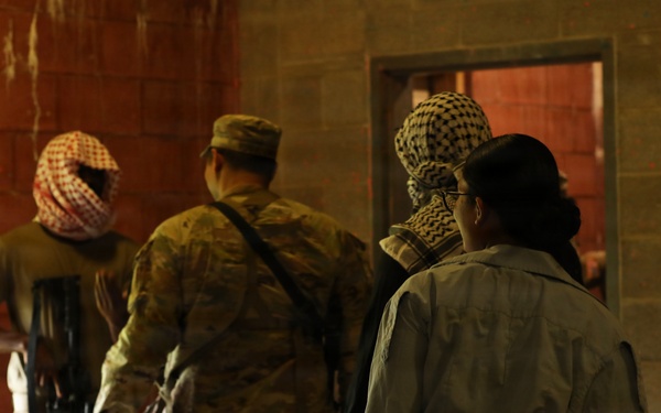 The image shows several individuals inside a brick building, viewed from behind. At least one person is wearing military camouflage uniform and a patrol cap, while others wear keffiyeh-style head coverings and mixed attire. The setting and equipment suggest a military training exercise or role-player scenario.