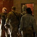The image shows several individuals inside a brick building, viewed from behind. At least one person is wearing military camouflage uniform and a patrol cap, while others wear keffiyeh-style head coverings and mixed attire. The setting and equipment suggest a military training exercise or role-player scenario.