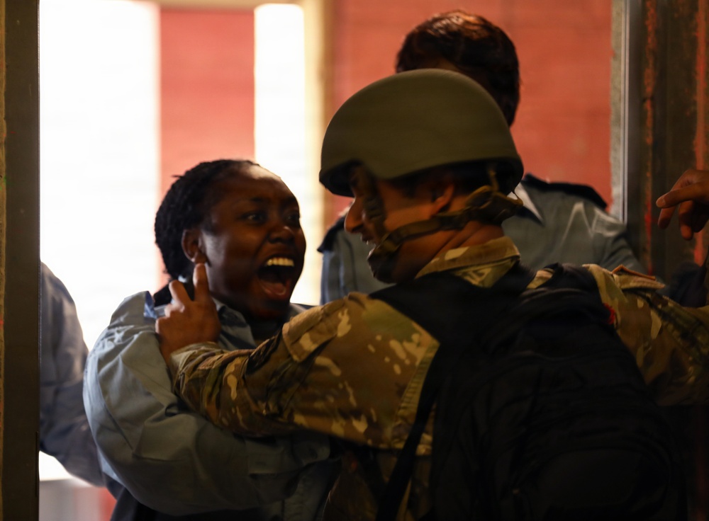 The image shows a tense interaction between a civilian woman and a soldier wearing military camouflage and a combat helmet. The woman appears distressed and is shouting, while the soldier appears to be restraining or guiding her. Additional individuals in blue uniforms, possibly police officers, are visible in the background.
