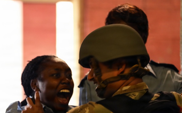 The image shows a tense interaction between a civilian woman and a soldier wearing military camouflage and a combat helmet. The woman appears distressed and is shouting, while the soldier appears to be restraining or guiding her. Additional individuals in blue uniforms, possibly police officers, are visible in the background.
