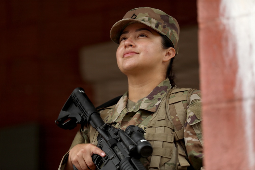 A female soldier in full military uniform is photographed from a low angle, gazing upward with a slight smile. She is wearing Operational Camouflage Pattern (OCP) uniform and patrol cap, holding an M4-style carbine rifle, and wearing a tactical vest. The image appears to be a professional or editorial military photograph taken near a brick structure.