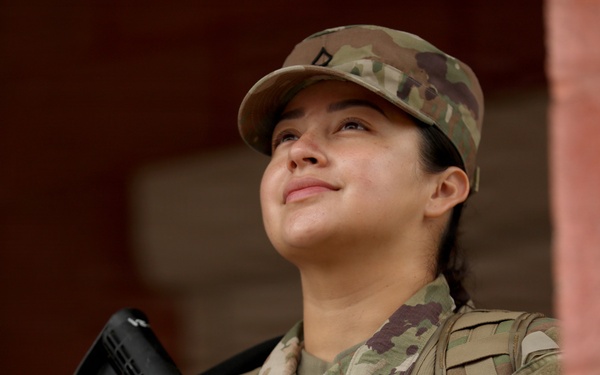 A female soldier in full military uniform is photographed from a low angle, gazing upward with a slight smile. She is wearing Operational Camouflage Pattern (OCP) uniform and patrol cap, holding an M4-style carbine rifle, and wearing a tactical vest. The image appears to be a professional or editorial military photograph taken near a brick structure.