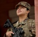 A female soldier in full military uniform is photographed from a low angle, gazing upward with a slight smile. She is wearing Operational Camouflage Pattern (OCP) uniform and patrol cap, holding an M4-style carbine rifle, and wearing a tactical vest. The image appears to be a professional or editorial military photograph taken near a brick structure.