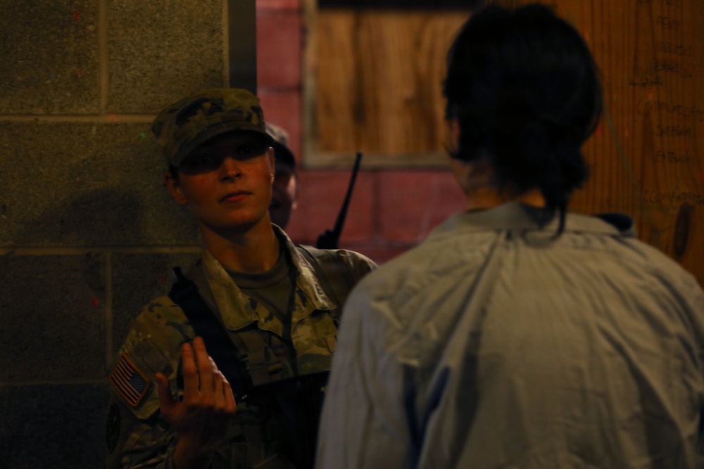 A U.S. Army soldier in MultiCam/OCP camouflage uniform is engaged in a face-to-face conversation with a civilian. The soldier is wearing a patrol cap, sunglasses, and a tactical vest with a radio visible, and has a U.S. flag patch on the left sleeve. The setting appears to be a dimly lit urban or training environment with concrete block walls.