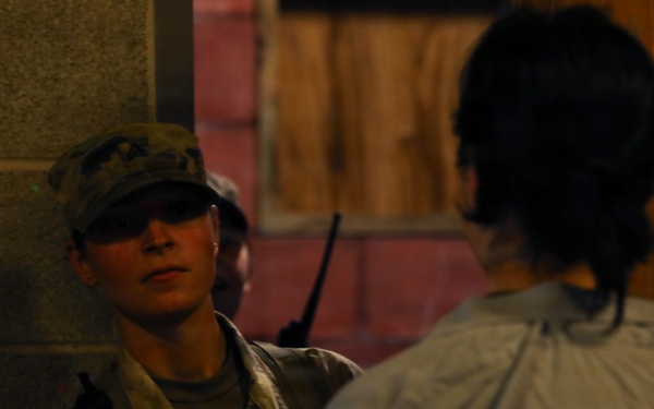 A U.S. Army soldier in MultiCam/OCP camouflage uniform is engaged in a face-to-face conversation with a civilian. The soldier is wearing a patrol cap, sunglasses, and a tactical vest with a radio visible, and has a U.S. flag patch on the left sleeve. The setting appears to be a dimly lit urban or training environment with concrete block walls.