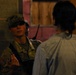 A U.S. Army soldier in MultiCam/OCP camouflage uniform is engaged in a face-to-face conversation with a civilian. The soldier is wearing a patrol cap, sunglasses, and a tactical vest with a radio visible, and has a U.S. flag patch on the left sleeve. The setting appears to be a dimly lit urban or training environment with concrete block walls.