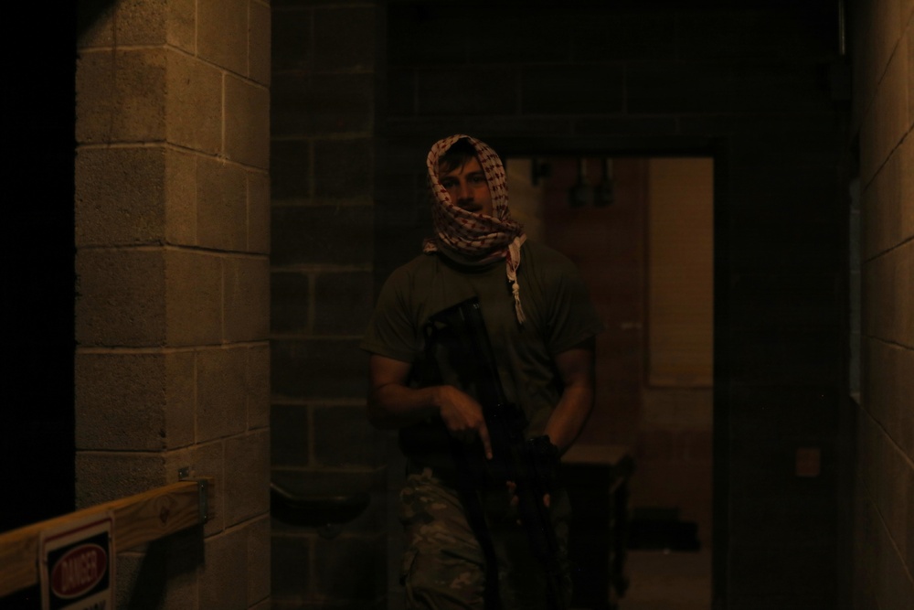 A person wearing a patterned head wrap (shemagh/keffiyeh) and camouflage pants is holding what appears to be a firearm or airsoft/training weapon in a dimly lit concrete block corridor or hallway. The setting appears to be an indoor training facility or simulated environment. The low lighting and tactical gear suggest a training exercise or staged scenario.