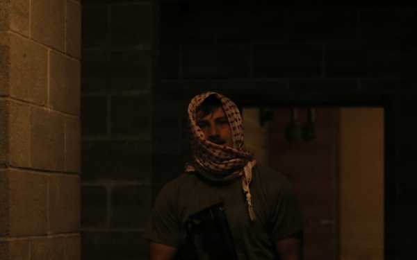 A person wearing a patterned head wrap (shemagh/keffiyeh) and camouflage pants is holding what appears to be a firearm or airsoft/training weapon in a dimly lit concrete block corridor or hallway. The setting appears to be an indoor training facility or simulated environment. The low lighting and tactical gear suggest a training exercise or staged scenario.