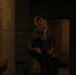 A person wearing a patterned head wrap (shemagh/keffiyeh) and camouflage pants is holding what appears to be a firearm or airsoft/training weapon in a dimly lit concrete block corridor or hallway. The setting appears to be an indoor training facility or simulated environment. The low lighting and tactical gear suggest a training exercise or staged scenario.