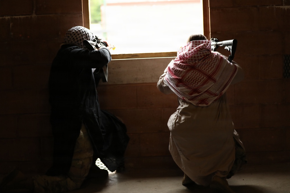 Two individuals are positioned inside a dark, brick-walled structure, aiming rifles through a window opening toward a brightly lit exterior. Both are wearing traditional Middle Eastern head coverings (keffiyeh). The image is taken from behind, showing the subjects in silhouette against the bright window.