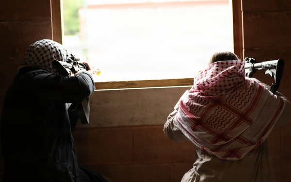 Two individuals are positioned inside a dark, brick-walled structure, aiming rifles through a window opening toward a brightly lit exterior. Both are wearing traditional Middle Eastern head coverings (keffiyeh). The image is taken from behind, showing the subjects in silhouette against the bright window.