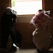 Two individuals are positioned inside a dark, brick-walled structure, aiming rifles through a window opening toward a brightly lit exterior. Both are wearing traditional Middle Eastern head coverings (keffiyeh). The image is taken from behind, showing the subjects in silhouette against the bright window.