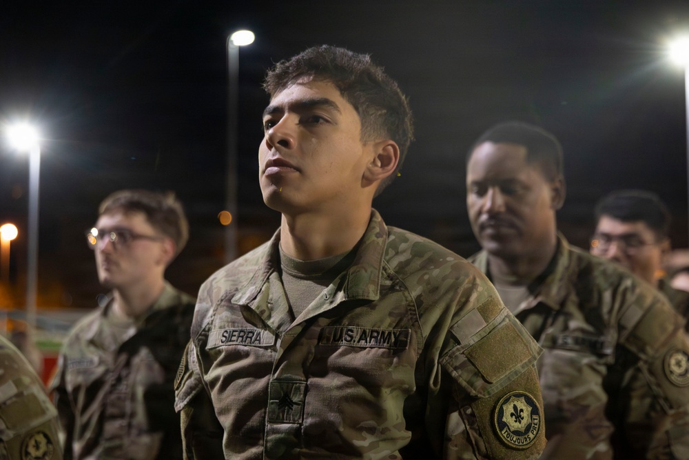 A group of U.S. Army soldiers in Operational Camouflage Pattern (OCP) uniforms are gathered outdoors at night under artificial lighting. The central figure, identified by name tape as 'SIERRA,' is standing at attention or listening attentively. A unit patch on the right sleeve reads 'Toujours Prêt,' associated with the 2nd Armored Cavalry Regiment.