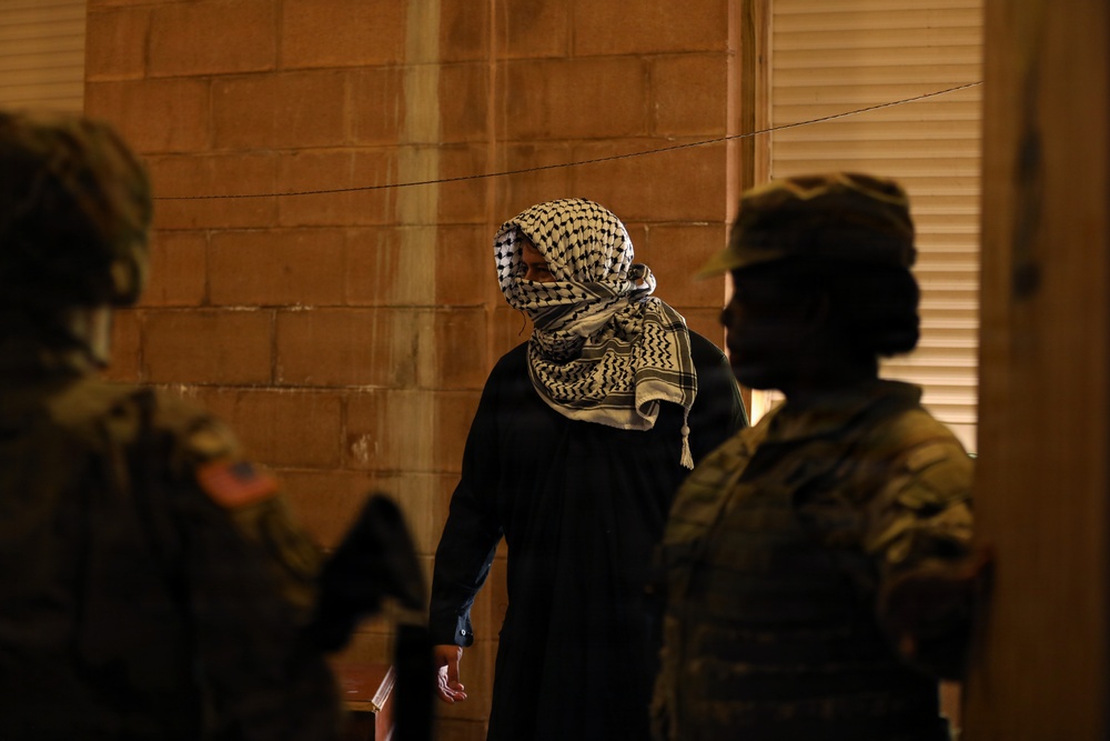 The image shows a nighttime scene with a central figure wearing a black outfit and a traditional keffiyeh/shemagh scarf covering their face, flanked by two individuals in military-style camouflage uniforms. The setting appears to be against a brick or concrete block wall with a window shutter visible on the right. The lighting is dim, creating strong shadows and silhouettes.