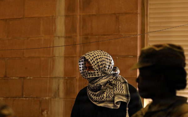 The image shows a nighttime scene with a central figure wearing a black outfit and a traditional keffiyeh/shemagh scarf covering their face, flanked by two individuals in military-style camouflage uniforms. The setting appears to be against a brick or concrete block wall with a window shutter visible on the right. The lighting is dim, creating strong shadows and silhouettes.