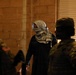 The image shows a nighttime scene with a central figure wearing a black outfit and a traditional keffiyeh/shemagh scarf covering their face, flanked by two individuals in military-style camouflage uniforms. The setting appears to be against a brick or concrete block wall with a window shutter visible on the right. The lighting is dim, creating strong shadows and silhouettes.
