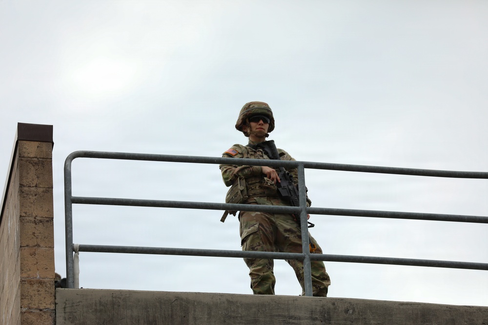 A uniformed military soldier stands on an elevated concrete structure, holding a rifle and leaning against a metal railing. The soldier is wearing full combat gear including a helmet, sunglasses, and camouflage uniform with a visible US flag patch on the left shoulder. The image is taken from a low angle looking upward against an overcast sky.