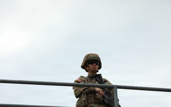 A uniformed military soldier stands on an elevated concrete structure, holding a rifle and leaning against a metal railing. The soldier is wearing full combat gear including a helmet, sunglasses, and camouflage uniform with a visible US flag patch on the left shoulder. The image is taken from a low angle looking upward against an overcast sky.