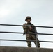 A uniformed military soldier stands on an elevated concrete structure, holding a rifle and leaning against a metal railing. The soldier is wearing full combat gear including a helmet, sunglasses, and camouflage uniform with a visible US flag patch on the left shoulder. The image is taken from a low angle looking upward against an overcast sky.