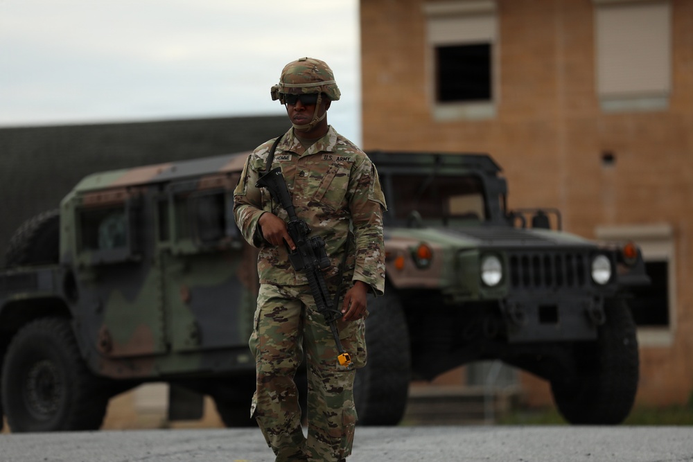 A U.S. Army soldier in Operational Camouflage Pattern (OCP) uniform is walking while carrying an assault rifle, with two military HMMWV (Humvee) vehicles visible in the background. The soldier is wearing a combat helmet, sunglasses, and has a name tape and U.S. Army tape visible on the uniform. The setting appears to be a military installation or training area with a brick building in the background.
