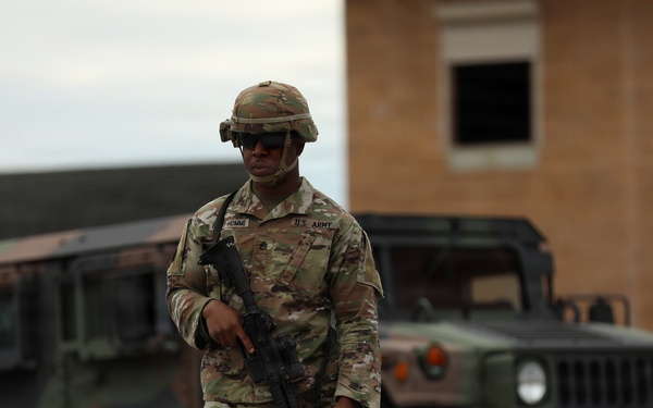 A U.S. Army soldier in Operational Camouflage Pattern (OCP) uniform is walking while carrying an assault rifle, with two military HMMWV (Humvee) vehicles visible in the background. The soldier is wearing a combat helmet, sunglasses, and has a name tape and U.S. Army tape visible on the uniform. The setting appears to be a military installation or training area with a brick building in the background.