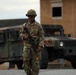 A U.S. Army soldier in Operational Camouflage Pattern (OCP) uniform is walking while carrying an assault rifle, with two military HMMWV (Humvee) vehicles visible in the background. The soldier is wearing a combat helmet, sunglasses, and has a name tape and U.S. Army tape visible on the uniform. The setting appears to be a military installation or training area with a brick building in the background.