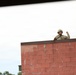 A soldier in camouflage uniform and helmet is visible above the roofline of a red brick building, with one fist raised in the air. The image is captured through a rectangular frame or window opening, giving it a framed-within-a-frame composition. The overcast sky provides a neutral background behind the figure.