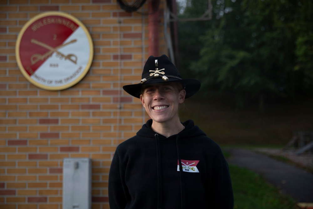 A smiling individual stands in front of a brick building wearing a black cowboy-style hat with crossed sabers insignia and a black hoodie with a unit patch. Behind them is a circular unit crest on the wall reading 'MULESKINNERS' with crossed cannons and the text 'STRENGTH OF THE DIVISION'. The setting appears to be a U.S. Army installation.