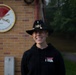 A smiling individual stands in front of a brick building wearing a black cowboy-style hat with crossed sabers insignia and a black hoodie with a unit patch. Behind them is a circular unit crest on the wall reading 'MULESKINNERS' with crossed cannons and the text 'STRENGTH OF THE DIVISION'. The setting appears to be a U.S. Army installation.
