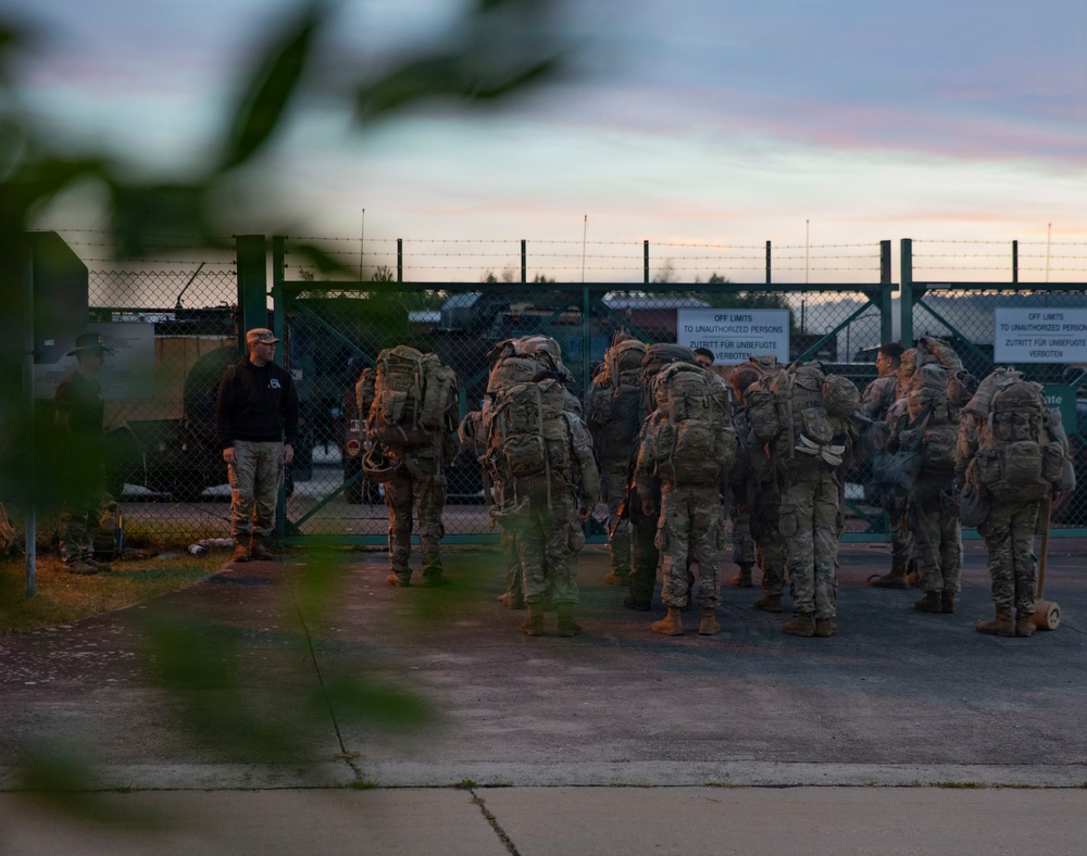 A group of military personnel in camouflage uniforms with large tactical backpacks are assembled near a secured gate at what appears to be a military installation, likely in Germany based on the bilingual German-English signage. The scene is photographed at dusk or dawn, with one individual in a black shirt standing apart from the uniformed group. The facility is surrounded by a chain-link fence topped with barbed wire.