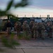 A group of military personnel in camouflage uniforms with large tactical backpacks are assembled near a secured gate at what appears to be a military installation, likely in Germany based on the bilingual German-English signage. The scene is photographed at dusk or dawn, with one individual in a black shirt standing apart from the uniformed group. The facility is surrounded by a chain-link fence topped with barbed wire.