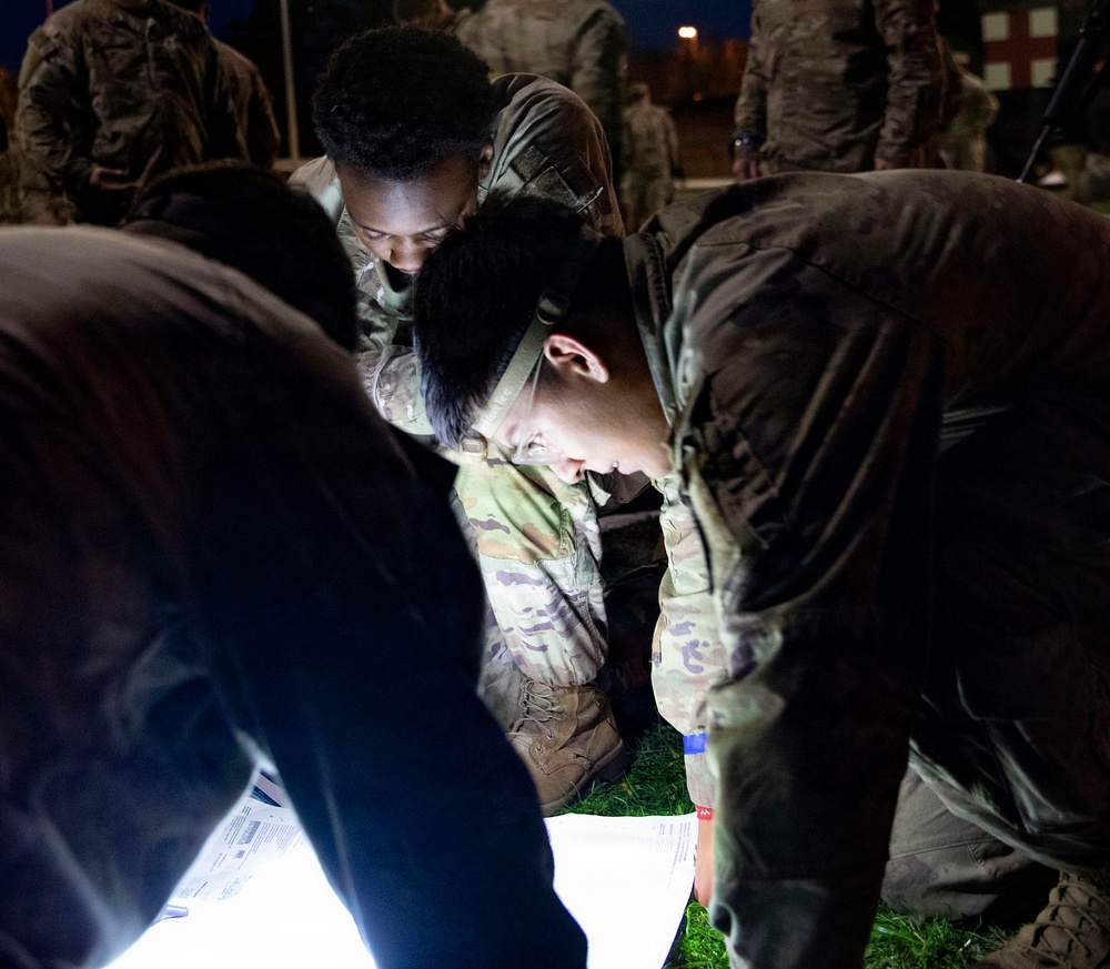 A group of military personnel in camouflage uniforms are gathered at night, huddled over illuminated documents on the ground. A headlamp worn by one soldier provides primary lighting for reading the papers. Additional soldiers are visible in the background near what appears to be a building or installation.