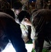 A group of military personnel in camouflage uniforms are gathered at night, huddled over illuminated documents on the ground. A headlamp worn by one soldier provides primary lighting for reading the papers. Additional soldiers are visible in the background near what appears to be a building or installation.