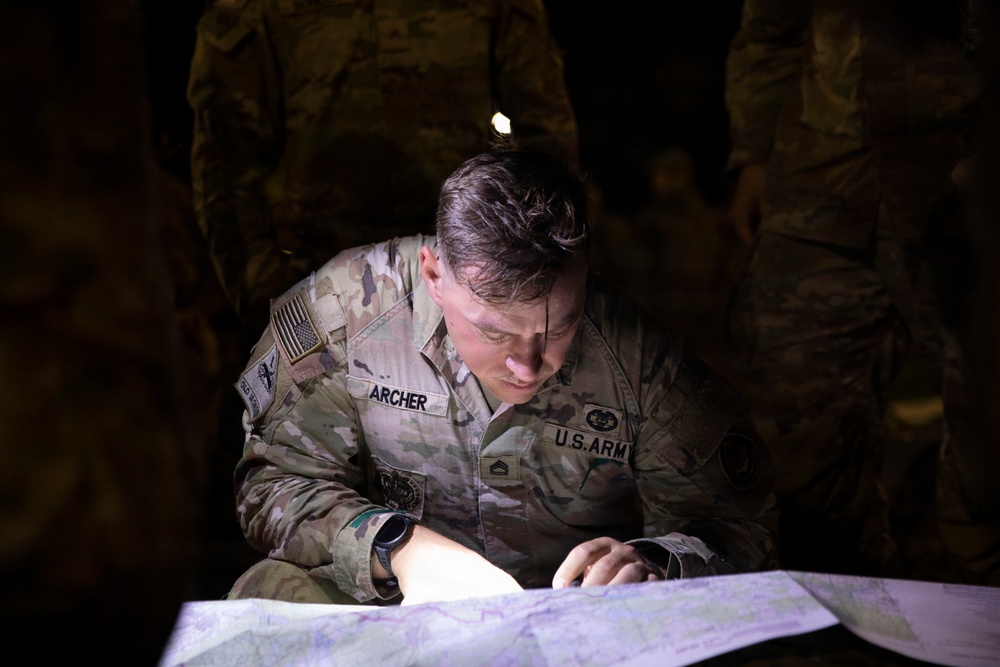 A U.S. Army soldier named Archer, identifiable by the name tape and branch tape on the uniform, is studying a topographic map in low-light conditions. The soldier appears to be conducting map reading or land navigation, illuminated by a focused light source. Additional soldiers in camouflage uniforms are visible in the dark background.
