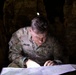A U.S. Army soldier named Archer, identifiable by the name tape and branch tape on the uniform, is studying a topographic map in low-light conditions. The soldier appears to be conducting map reading or land navigation, illuminated by a focused light source. Additional soldiers in camouflage uniforms are visible in the dark background.