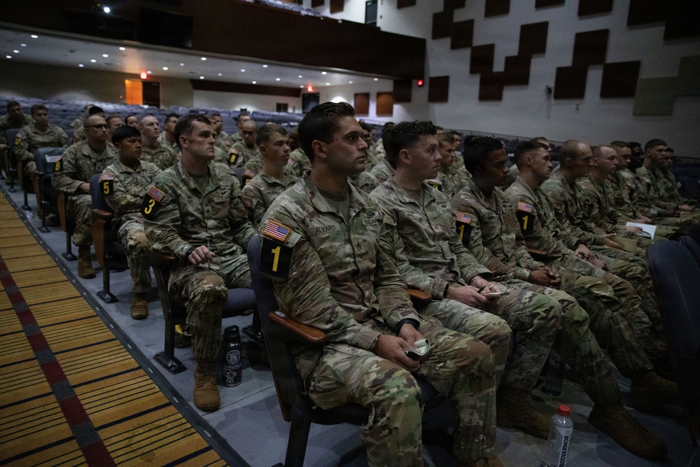 A large group of U.S. Army soldiers in MultiCam/OCP camouflage uniforms are seated in an auditorium or theater-style briefing room. The soldiers are arranged in numbered rows (visible number placards 1, 3, 5, 8) and appear to be attending a formal briefing or presentation. American flag patches are visible on their uniforms, and one soldier's name tape reads 'ALVARO'.