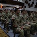 A large group of U.S. Army soldiers in MultiCam/OCP camouflage uniforms are seated in an auditorium or theater-style briefing room. The soldiers are arranged in numbered rows (visible number placards 1, 3, 5, 8) and appear to be attending a formal briefing or presentation. American flag patches are visible on their uniforms, and one soldier's name tape reads 'ALVARO'.