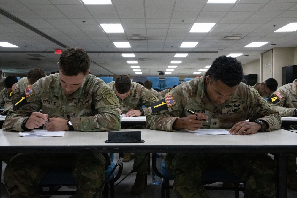 Multiple U.S. Army soldiers in OCP (Operational Camouflage Pattern) uniforms are seated at tables in a classroom or testing facility, writing on papers. The two foreground soldiers are identified by name tapes as 'VERETTE' and 'JOHNSON', both wearing U.S. Army insignia and American flag patches. The setting appears to be a formal written examination or administrative processing event.