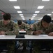 Multiple U.S. Army soldiers in OCP (Operational Camouflage Pattern) uniforms are seated at tables in a classroom or testing facility, writing on papers. The two foreground soldiers are identified by name tapes as 'VERETTE' and 'JOHNSON', both wearing U.S. Army insignia and American flag patches. The setting appears to be a formal written examination or administrative processing event.