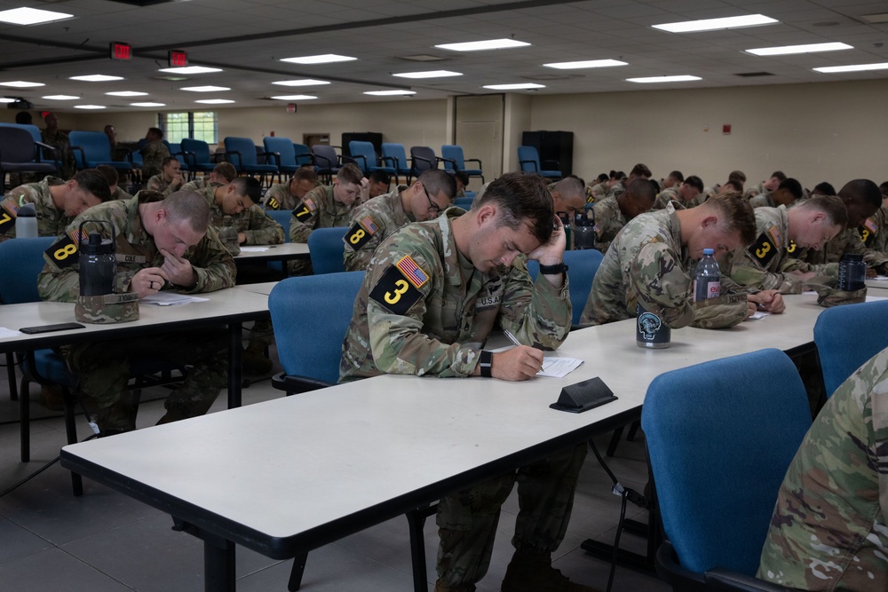 A large group of U.S. Army soldiers in OCP (Operational Camouflage Pattern) uniforms are seated at tables in a classroom or conference room, actively writing on papers. The soldiers are wearing numbered placards (visible numbers include 3, 7, 8) on their uniforms, suggesting a competition or evaluation event. The setting appears to be a military training facility with fluorescent lighting and blue chairs.