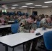 A large group of U.S. Army soldiers in OCP (Operational Camouflage Pattern) uniforms are seated at tables in a classroom or conference room, actively writing on papers. The soldiers are wearing numbered placards (visible numbers include 3, 7, 8) on their uniforms, suggesting a competition or evaluation event. The setting appears to be a military training facility with fluorescent lighting and blue chairs.