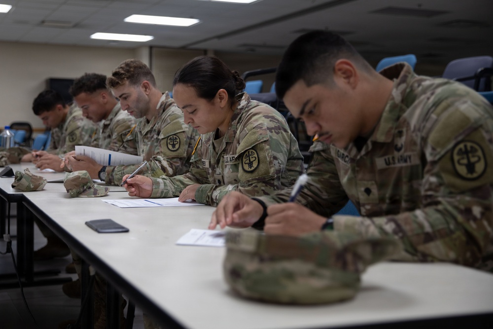 A group of U.S. Army soldiers in Operational Camouflage Pattern (OCP) uniforms are seated at a long white table, writing on papers in what appears to be a classroom or briefing room setting. The soldiers display shoulder patches consistent with a medical unit (caduceus-style insignia). The scene suggests a written examination, training exercise, or administrative paperwork session.