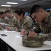 A group of U.S. Army soldiers in Operational Camouflage Pattern (OCP) uniforms are seated at a long white table, writing on papers in what appears to be a classroom or briefing room setting. The soldiers display shoulder patches consistent with a medical unit (caduceus-style insignia). The scene suggests a written examination, training exercise, or administrative paperwork session.