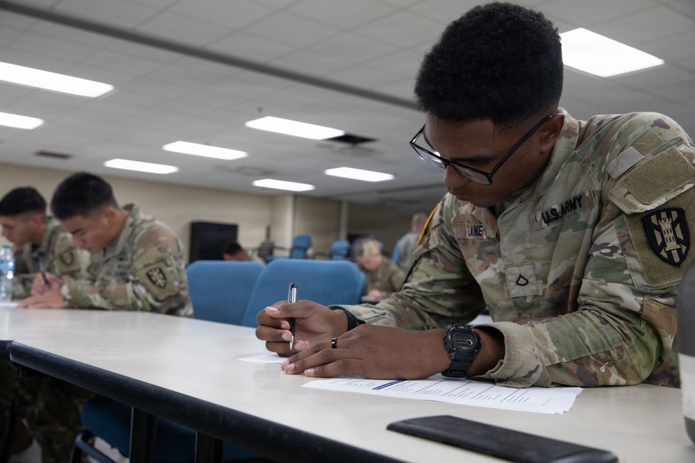 A U.S. Army soldier wearing OCP (Operational Camouflage Pattern) uniform is seated at a table writing on a document with a pen. The setting appears to be a military classroom or briefing room with multiple other soldiers visible in the background doing similar tasks. The foreground soldier is wearing glasses, a watch, and has a name tape reading 'LANE' with a Private First Class rank insignia visible.