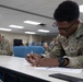 A U.S. Army soldier wearing OCP (Operational Camouflage Pattern) uniform is seated at a table writing on a document with a pen. The setting appears to be a military classroom or briefing room with multiple other soldiers visible in the background doing similar tasks. The foreground soldier is wearing glasses, a watch, and has a name tape reading 'LANE' with a Private First Class rank insignia visible.