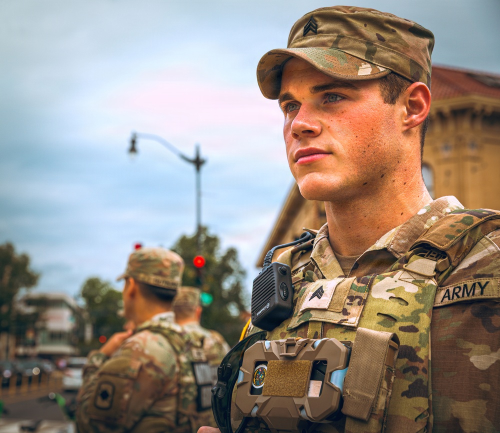 A U.S. Army soldier in the foreground wearing Operational Camouflage Pattern (OCP) uniform and body armor is standing alert in an urban environment. He is equipped with a radio/communications device clipped to his vest and displays Sergeant rank insignia on both his cap and chest. At least two additional soldiers are visible in the background near a traffic intersection.