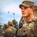 A U.S. Army soldier in the foreground wearing Operational Camouflage Pattern (OCP) uniform and body armor is standing alert in an urban environment. He is equipped with a radio/communications device clipped to his vest and displays Sergeant rank insignia on both his cap and chest. At least two additional soldiers are visible in the background near a traffic intersection.