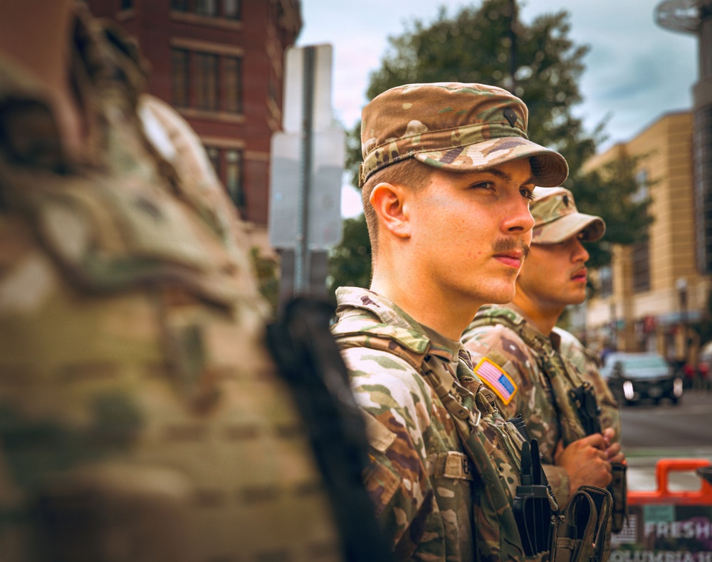 The image shows at least three U.S. military personnel in MultiCam (OCP) camouflage uniforms standing in formation on an urban street. The soldiers are wearing tactical gear including plate carriers and are equipped with weapons. They appear to be on a domestic deployment or security detail in a city environment.