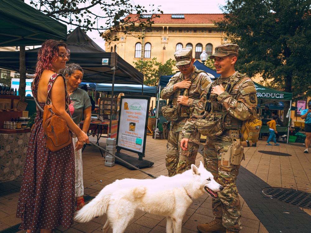 Two U.S. Army soldiers in full camouflage uniforms and tactical gear are interacting with civilians at an outdoor farmers market or street fair. A woman with pink hair and a flowing dress holds a white dog on a leash while conversing with the soldiers. The scene takes place in an urban plaza surrounded by vendor tents and a historic brick building in the background.